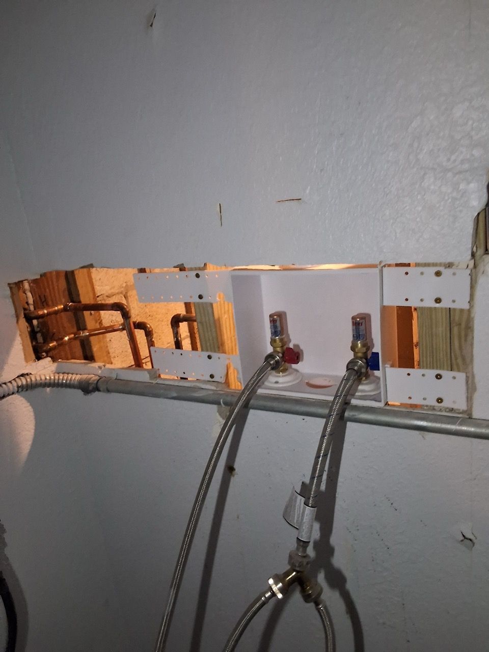Wall opening with exposed plumbing valves and braided hoses in a gray interior wall