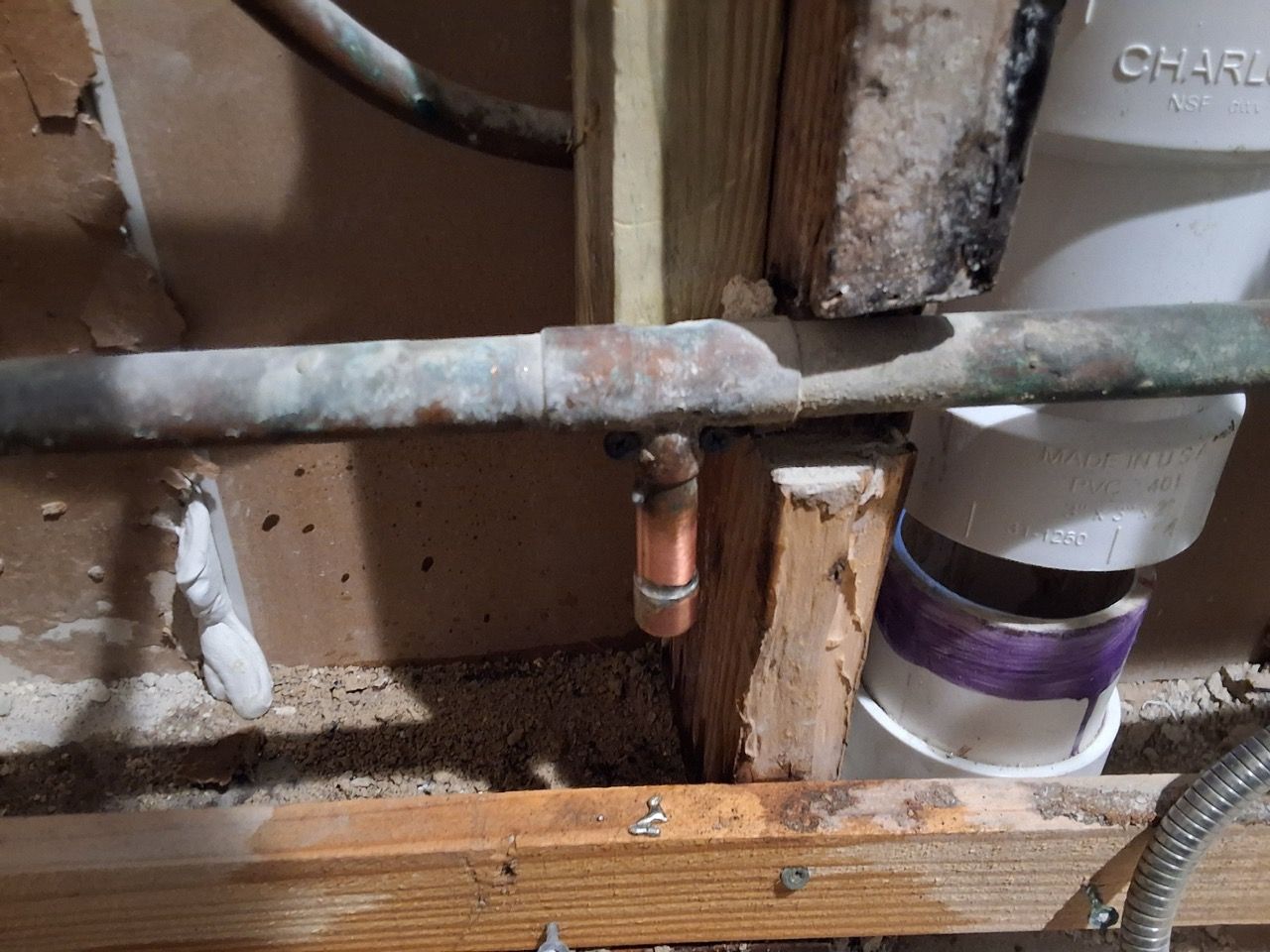 Under-sink plumbing with a corroded copper pipe, wooden framing, and a purple banded container on the right