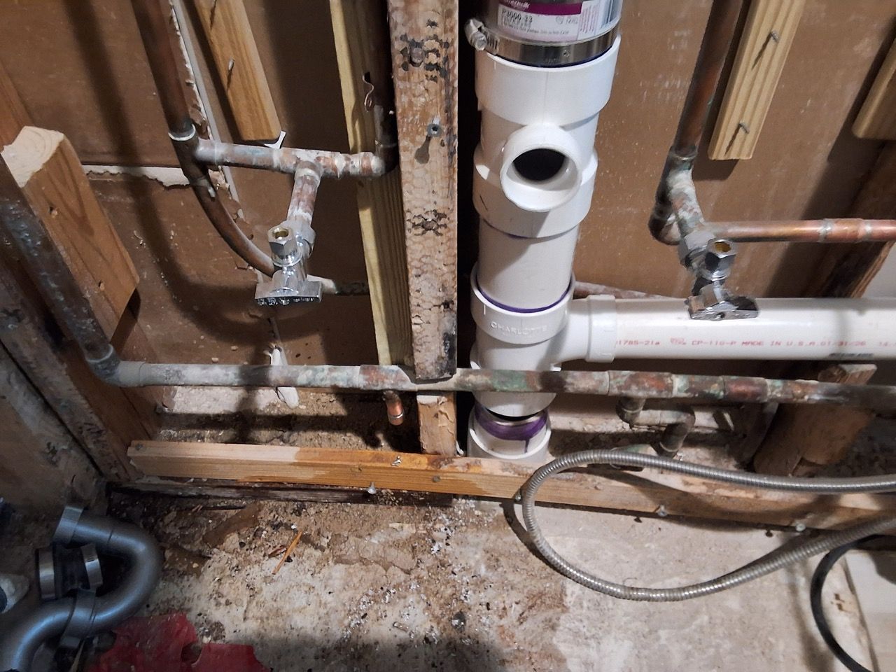 Exposed plumbing pipes and valves under a sink, with damp floor and damaged wood framing.