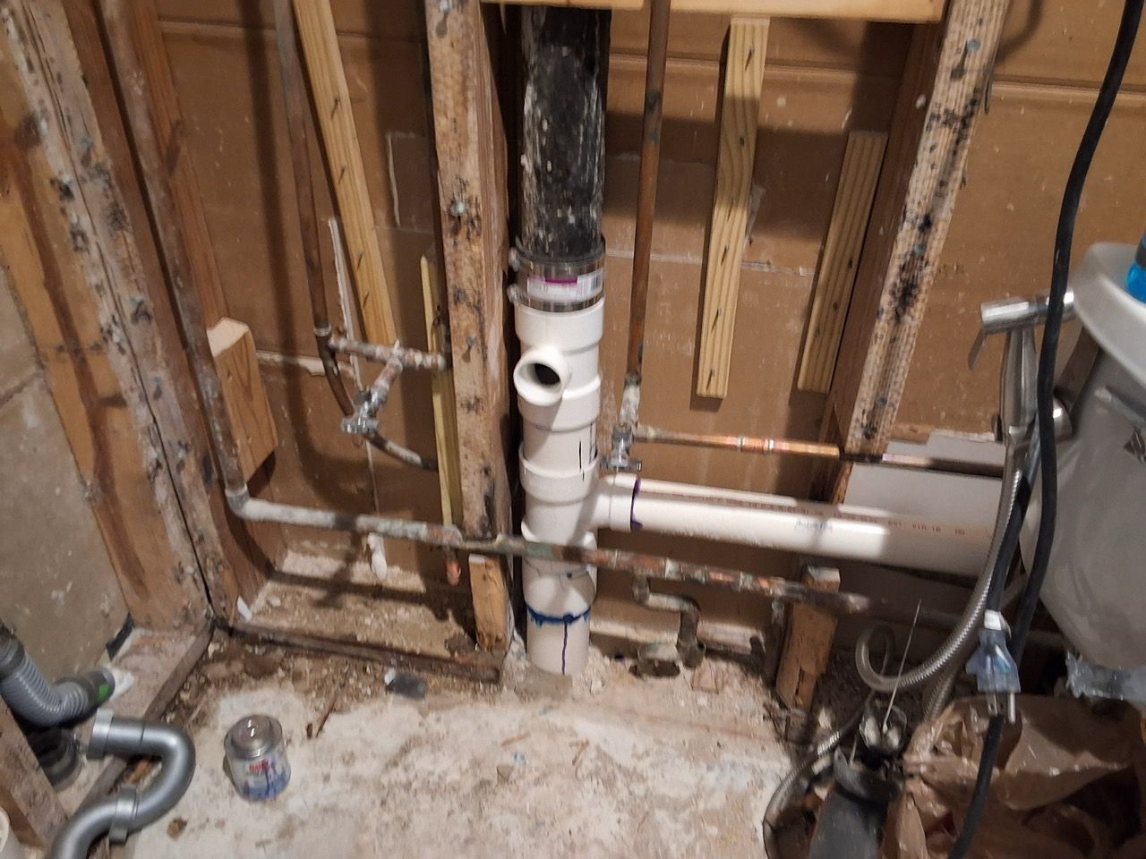 Exposed plumbing and PVC pipes in an unfinished bathroom wall with studs and debris on the floor