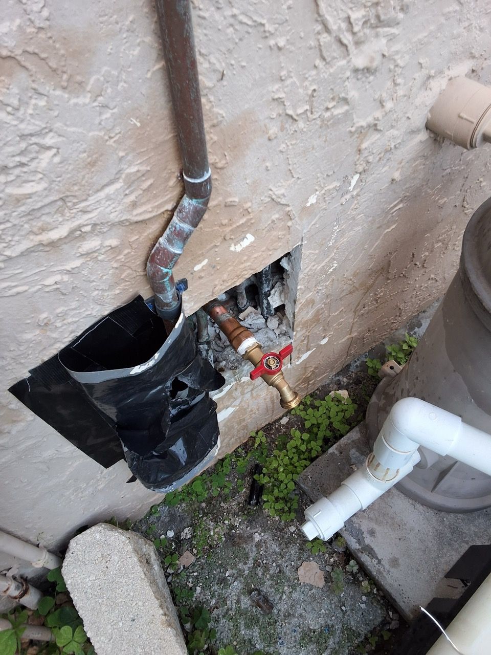 Damaged wall with exposed black ducting and pipes beside a utility meter and white plumbing pipe.