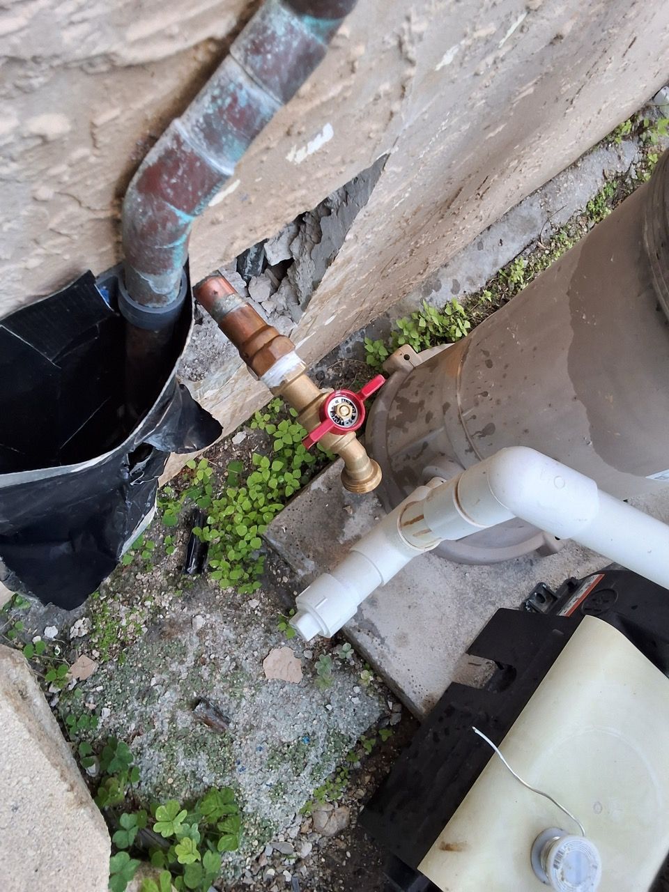 Outdoor plumbing pipes and a red shutoff valve beside a wall, with weeds and gravel below.