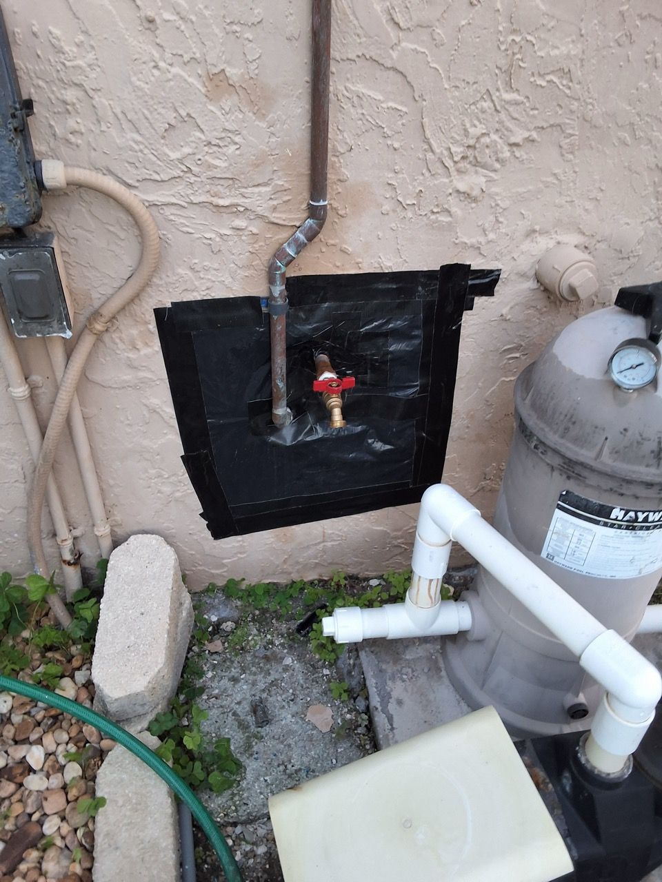 Outdoor utility area with gas meter, exposed pipes, and a black wall patch.
