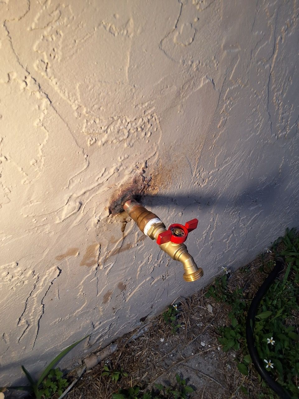 Outdoor wall-mounted faucet with a red handle and a pipe extending from a stucco wall, beside dirt and grass.