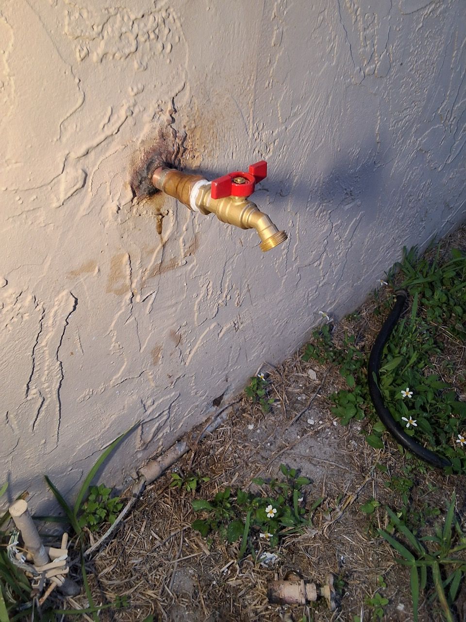 Rusty outdoor faucet on a beige wall beside grass and dirt.