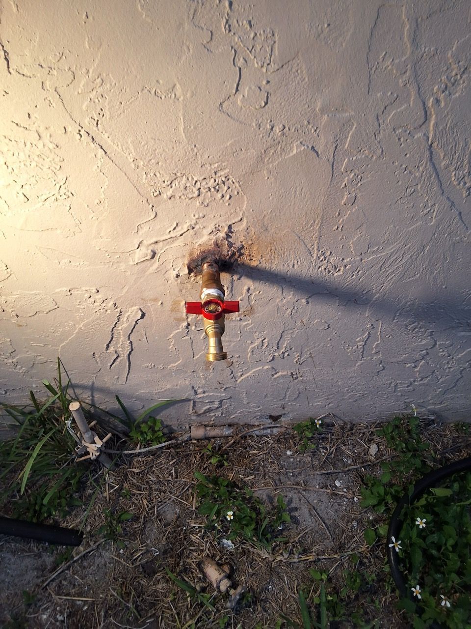 Outdoor wall faucet with a red valve and yellow handle on a stucco wall near grass and dirt.