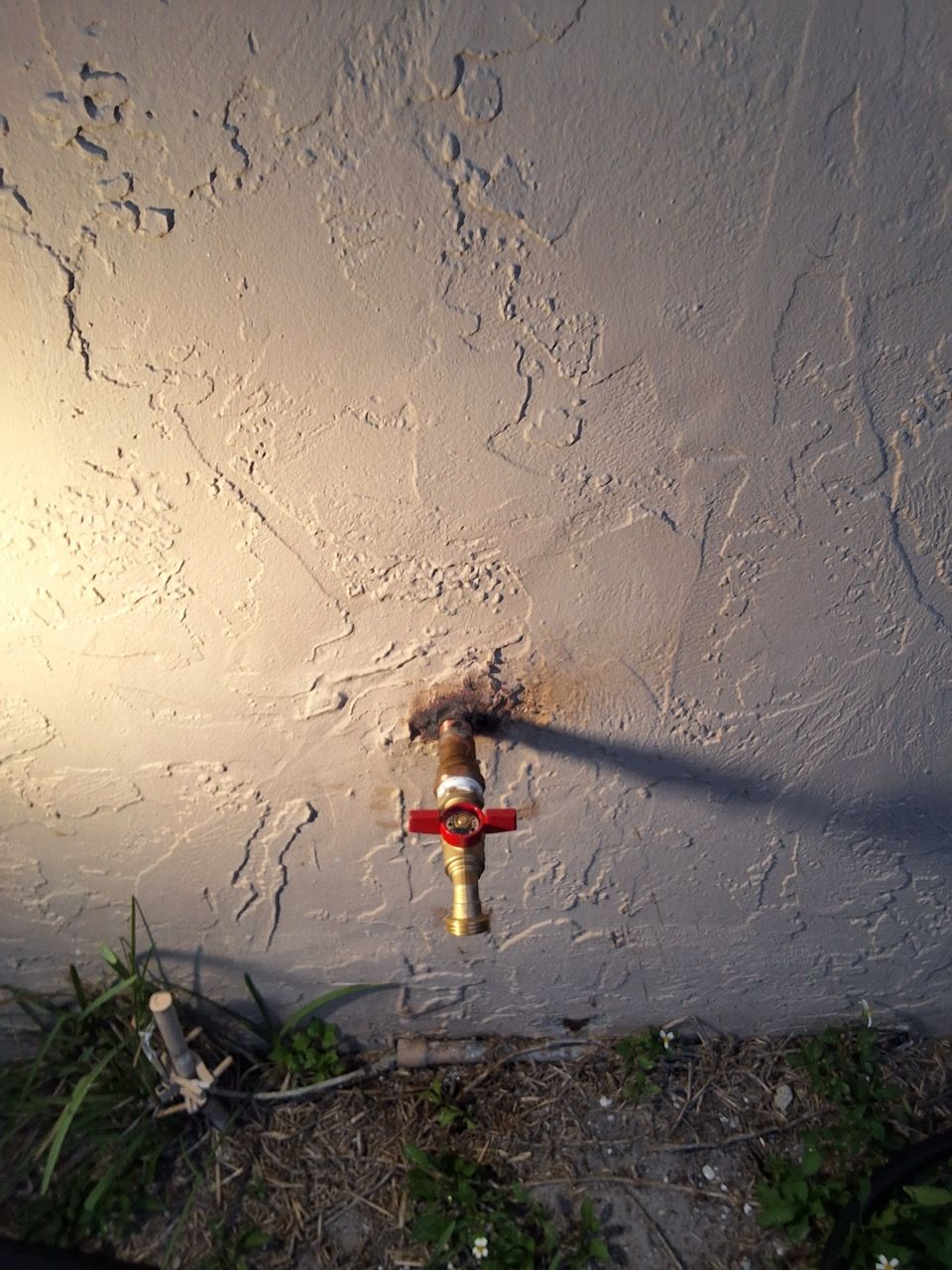 Outdoor wall faucet with red handle and yellow spout, mounted on a stucco wall above dirt and grass.