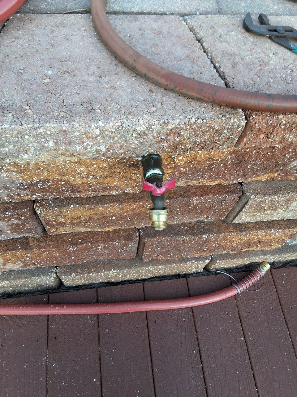 Outdoor hose spigot mounted on a brick wall above a wooden deck, with red hose attached.