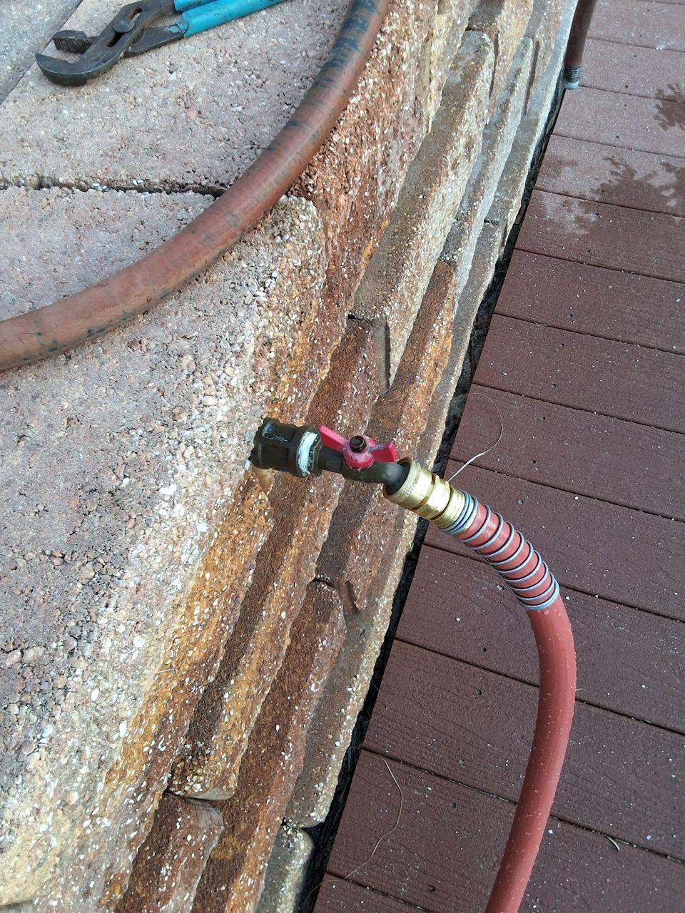 Coiled hose with metal nozzle beside a weathered wooden deck and concrete curb.
