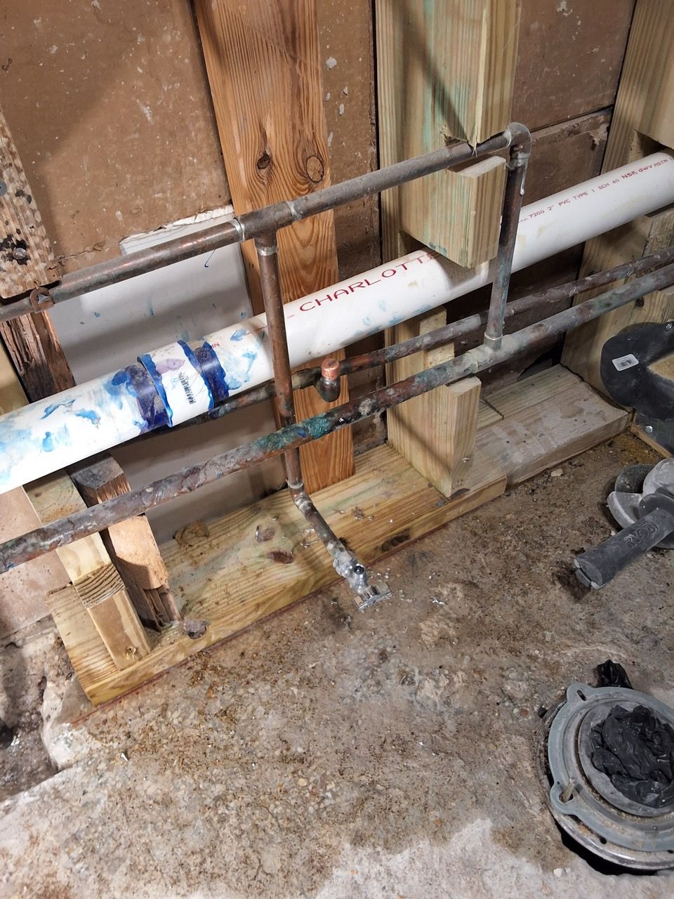 Black pipe clamped to wood supports in a utility area, with a white PVC pipe and floor debris nearby