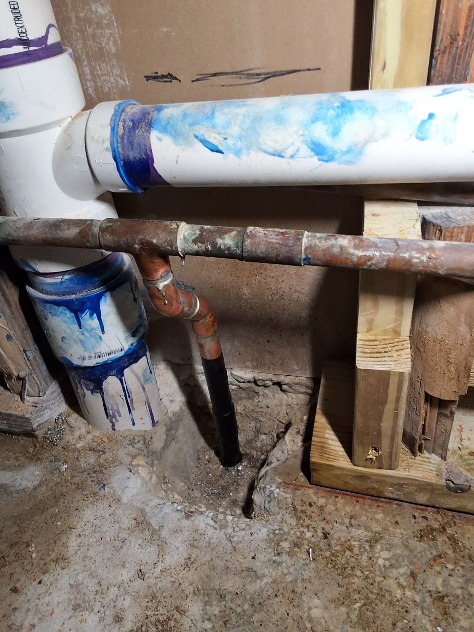 Leaking white PVC pipe with blue stains above a wet concrete floor and wooden supports