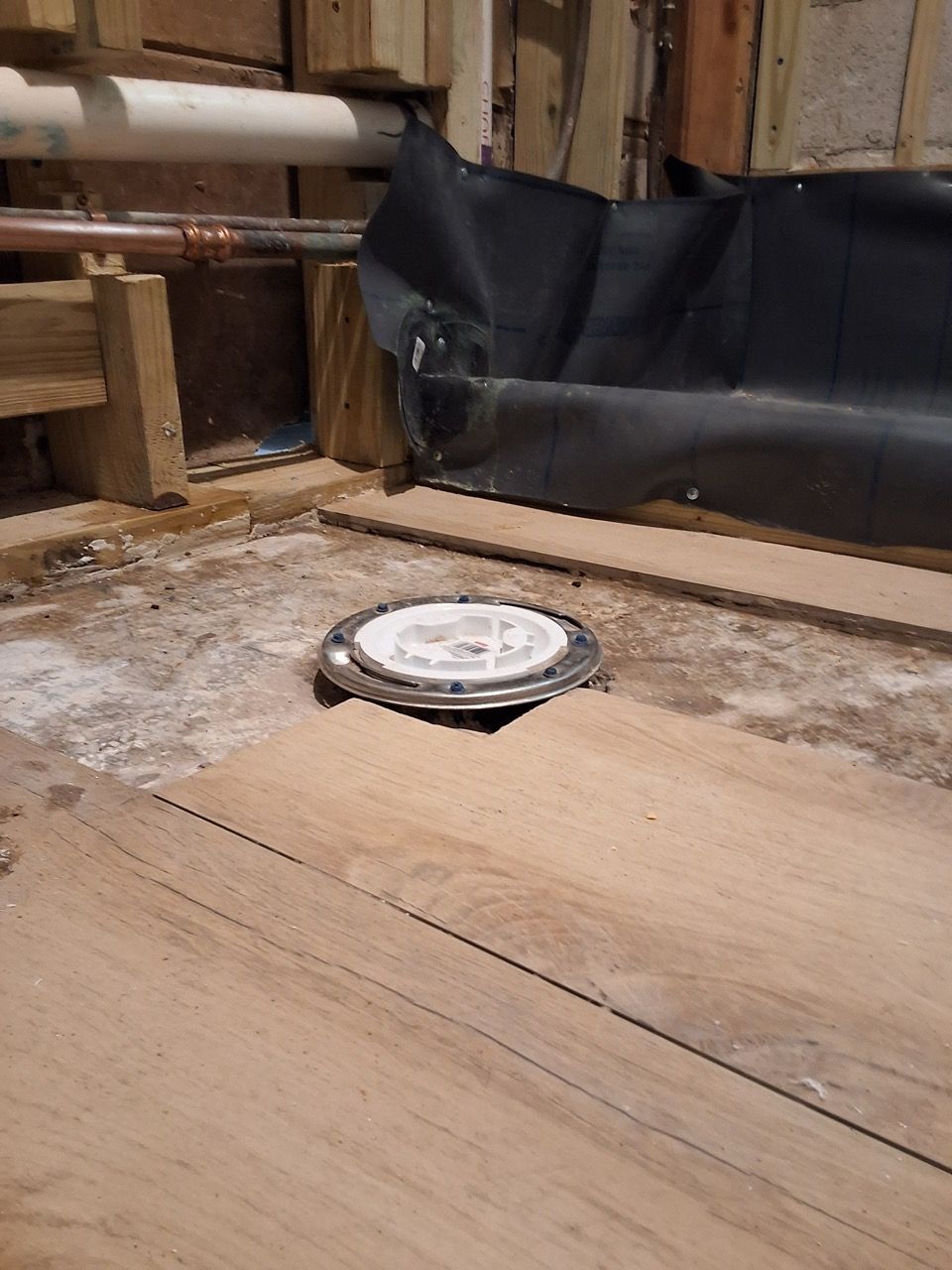Round metal sensor embedded in a dusty wooden floor beside black plastic sheeting and exposed pipes.