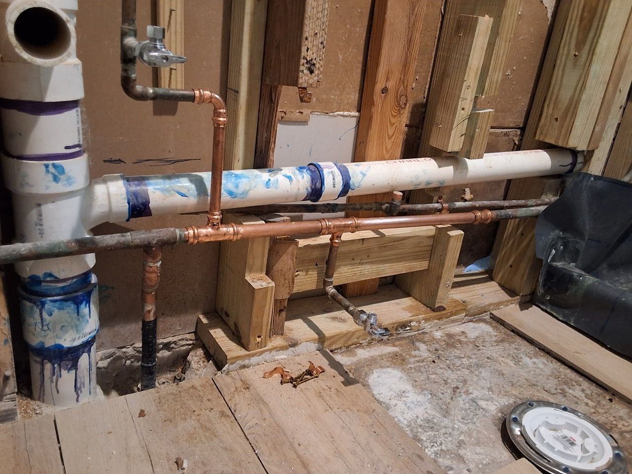 Exposed plumbing pipes and valves inside an unfinished wall framing area with debris on the floor.