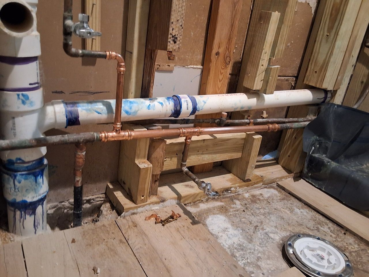 Exposed plumbing pipes and fittings in an unfinished room with wooden framing and debris on the floor
