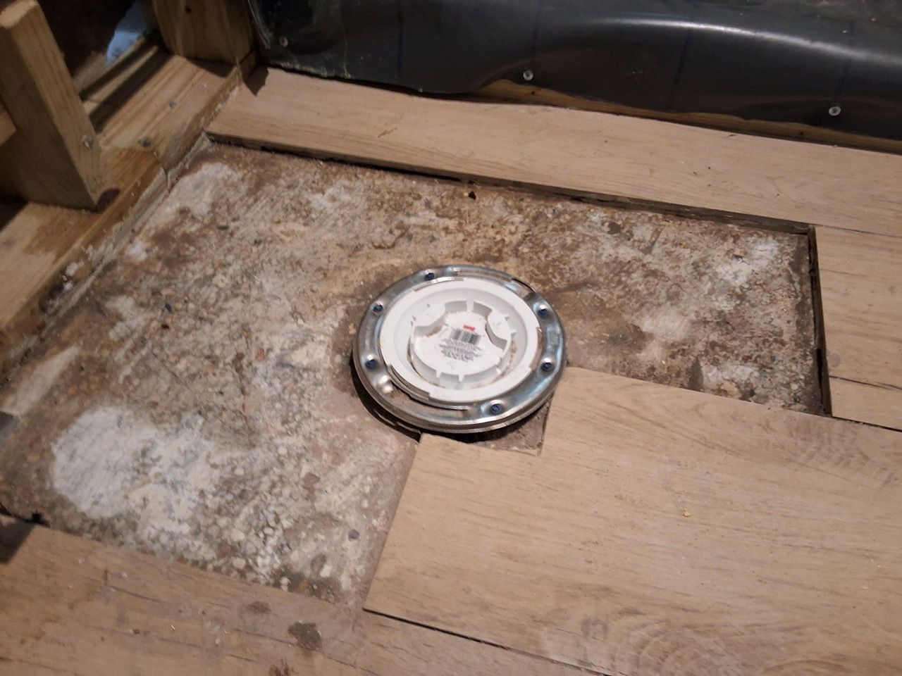 Round floor drain in a dusty wooden floor opening, surrounded by plywood and debris.