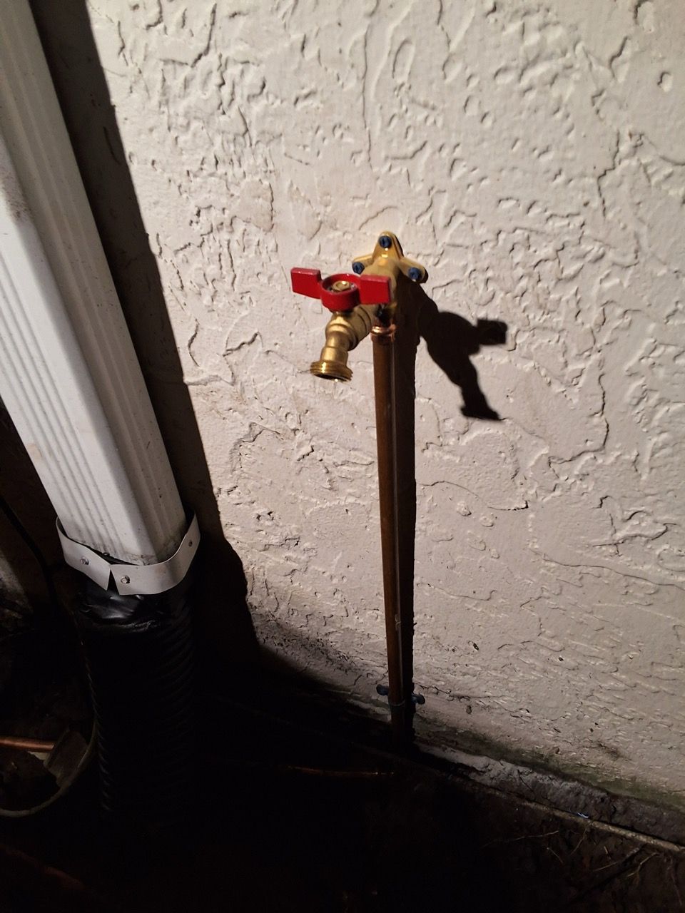 Outdoor water spigot with red handle mounted on a wall beside a drainpipe at night