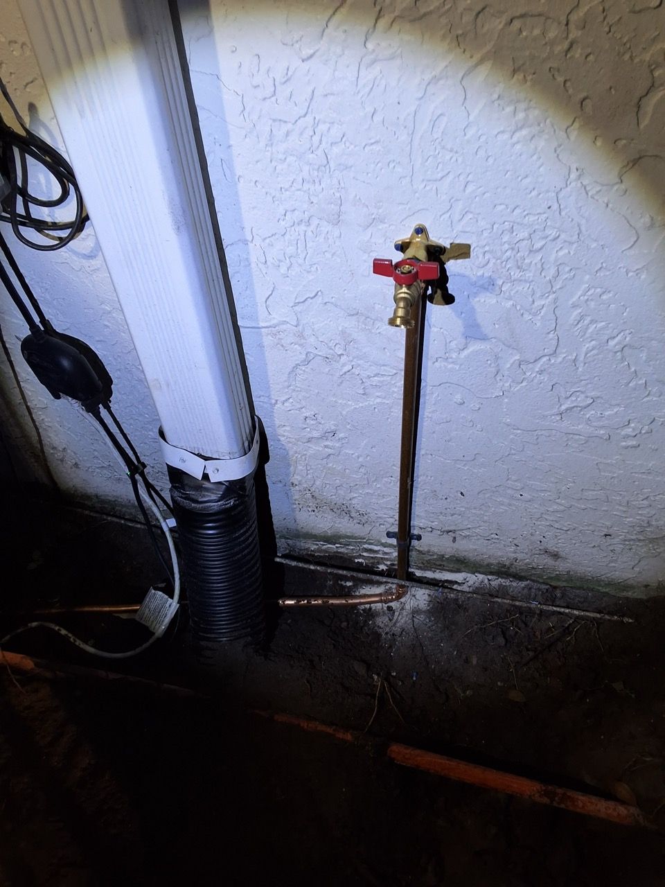 Wall-mounted outdoor faucet with red handle beside a white pipe on a stucco wall at night.