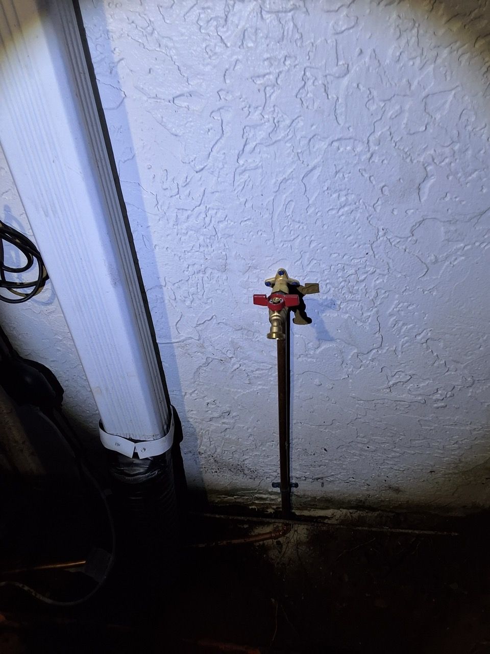 Outdoor water spigot on a stucco wall beside a downspout, with a small valve handle and dark ground below