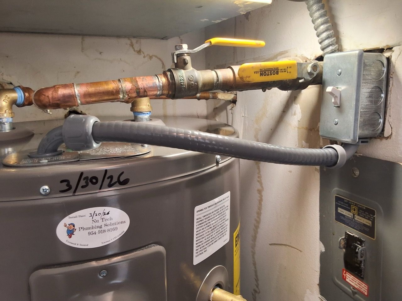 Water heater with copper and black gas pipes, shutoff valve, and control box in a utility room