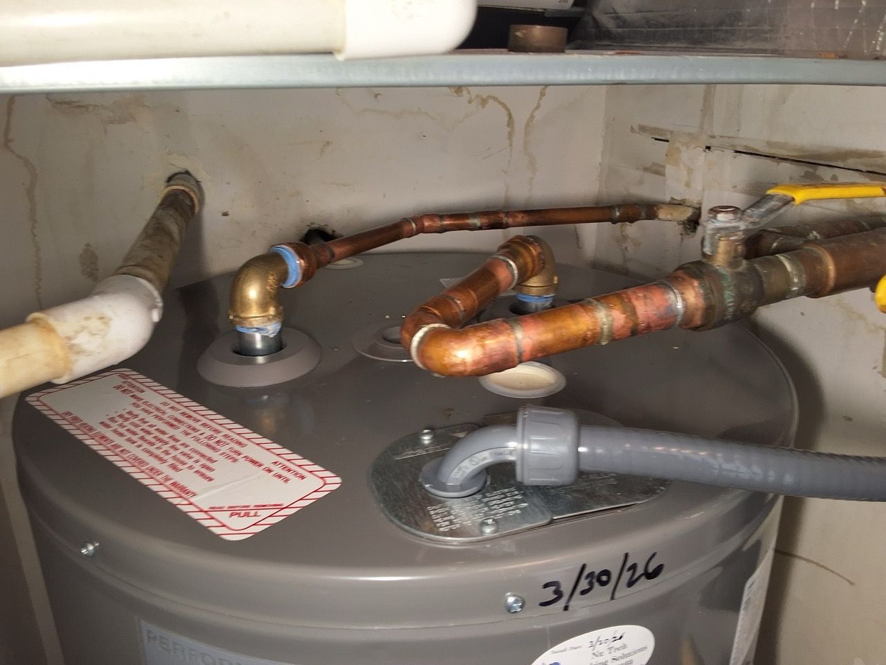 Water heater with exposed copper pipes and gray hoses in a utility cabinet, showing minor rust and labels.