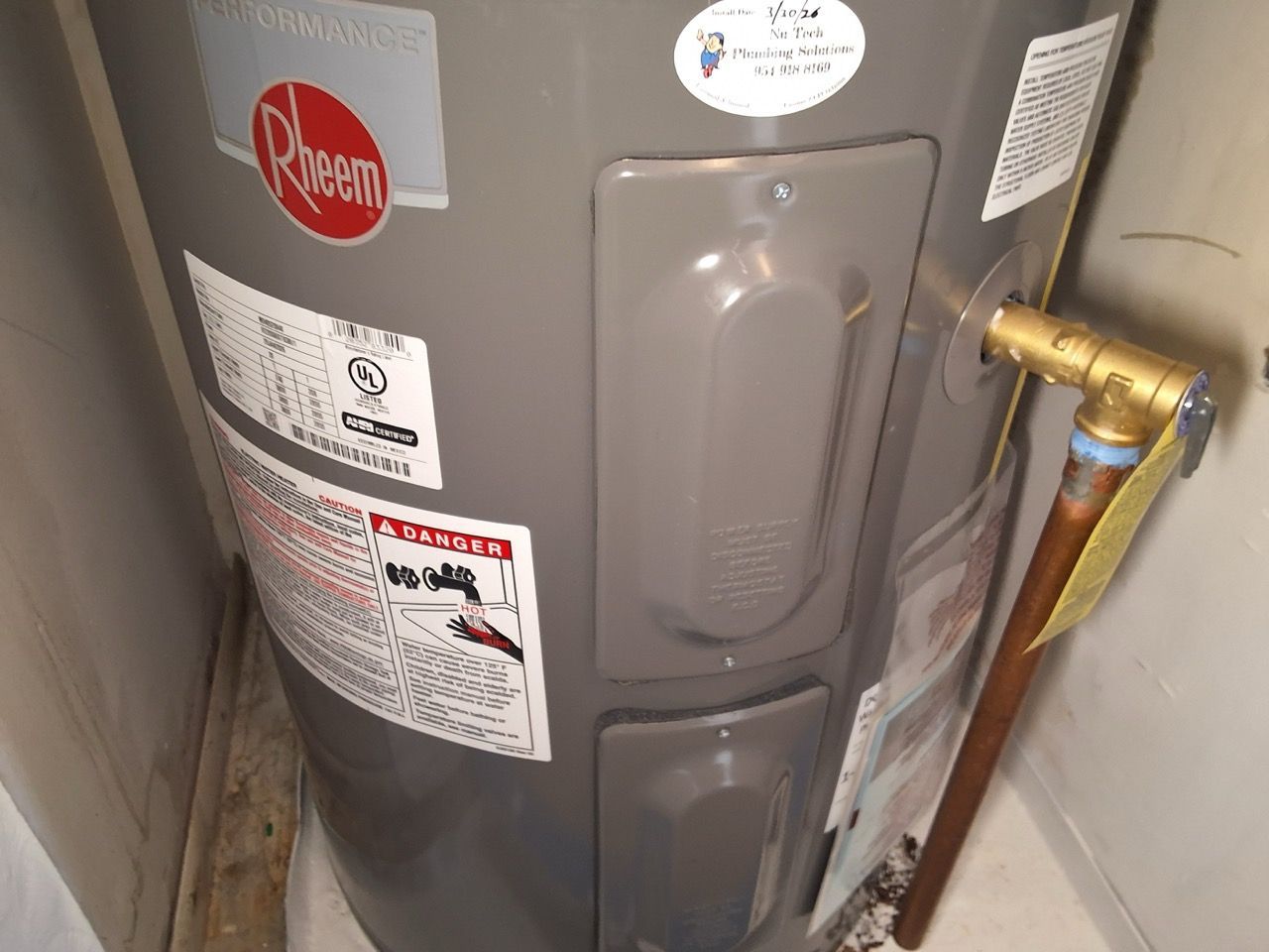Gray Rheem water heater with brass plumbing and labels in a utility closet