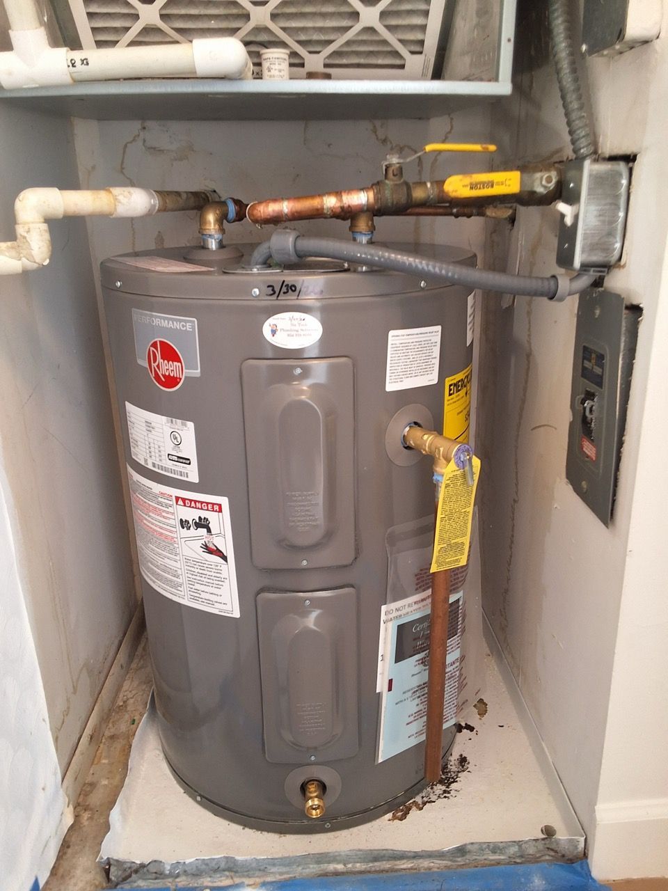 Gray water heater in a utility closet with pipes, valves, and labels attached.
