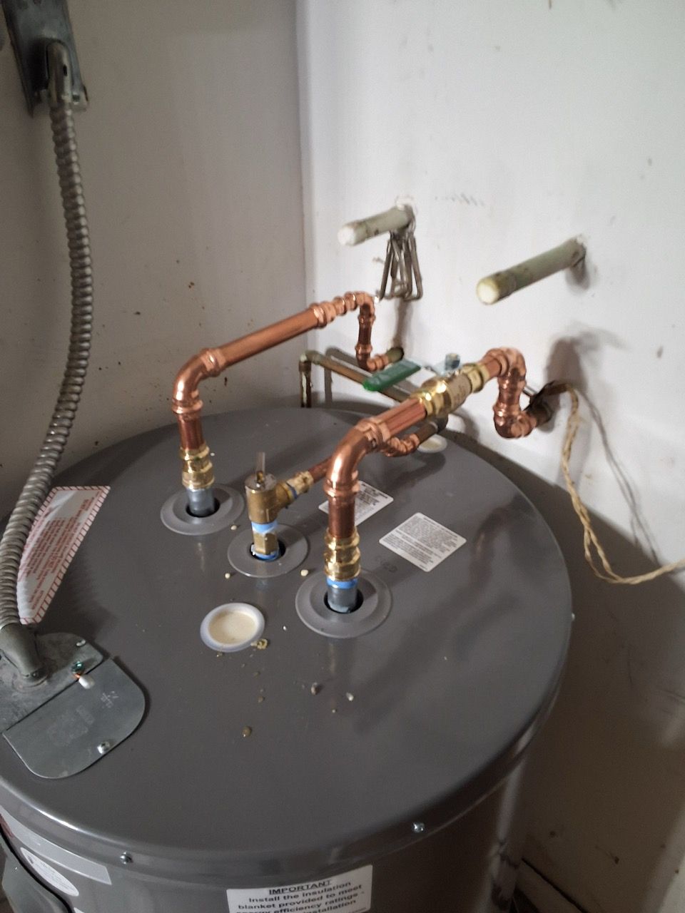 Water heater with copper pipes and valves in a utility corner against a wall.