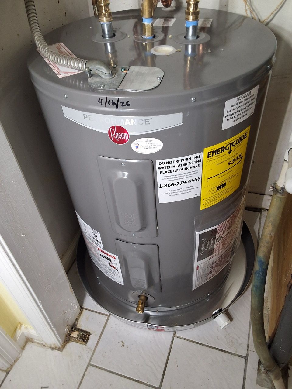 Gray water heater in a utility closet with warning labels and pipes on top, beside white tile floor.