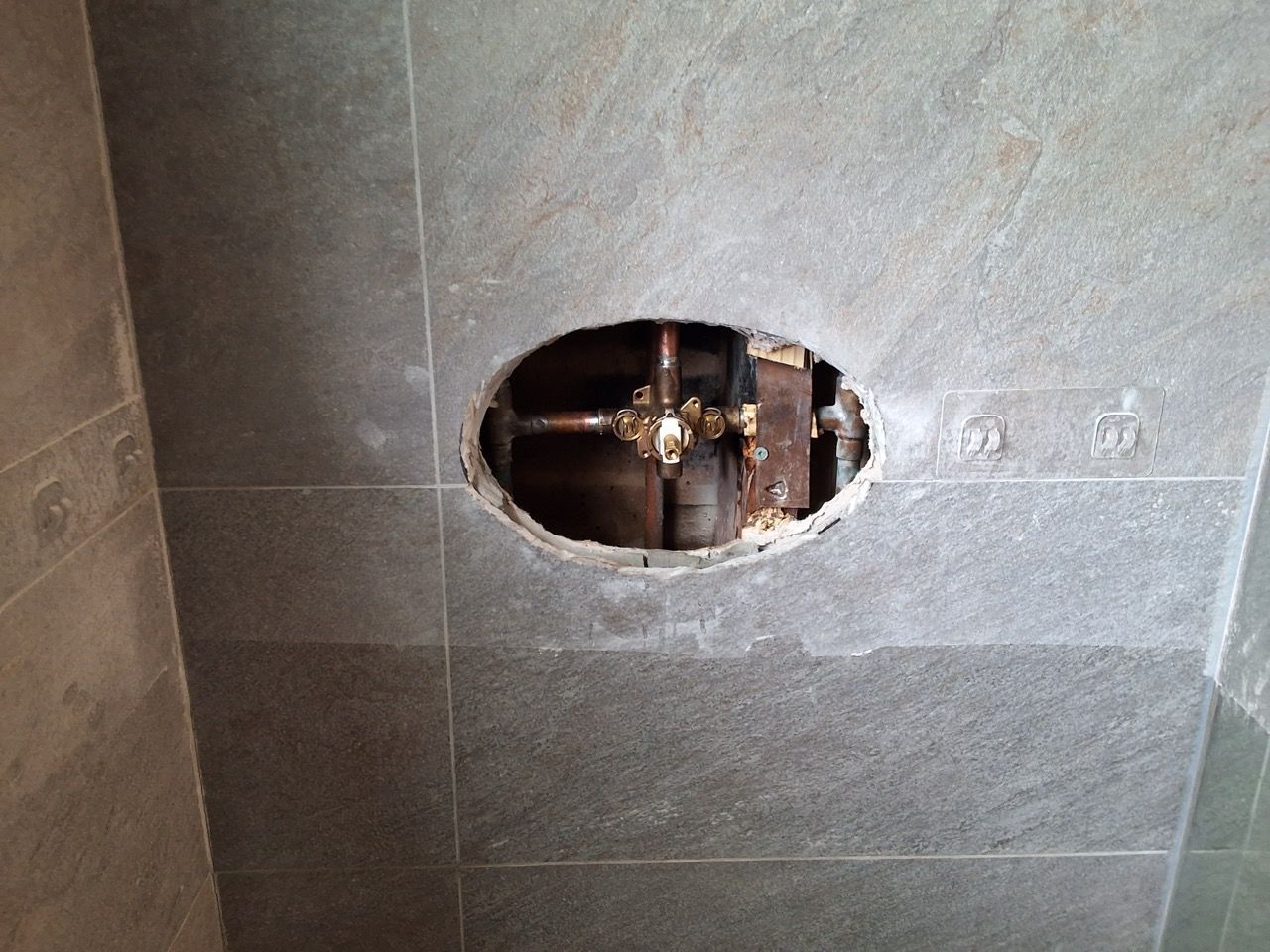 Gray tiled wall with an oval hole exposing plumbing pipes and valves inside.