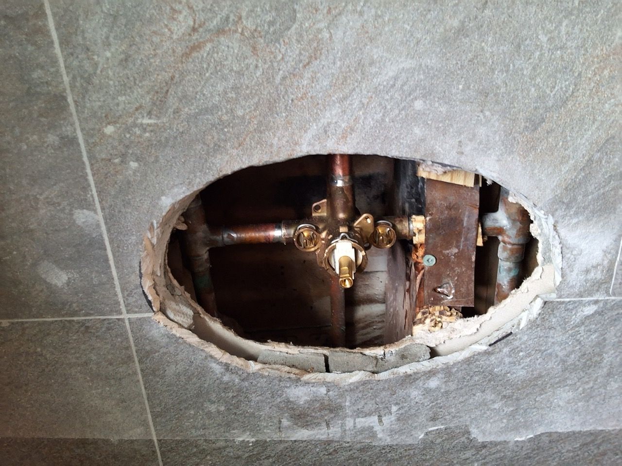 Broken wall exposing exposed copper pipes and valve behind a rough oval hole in tiled surface