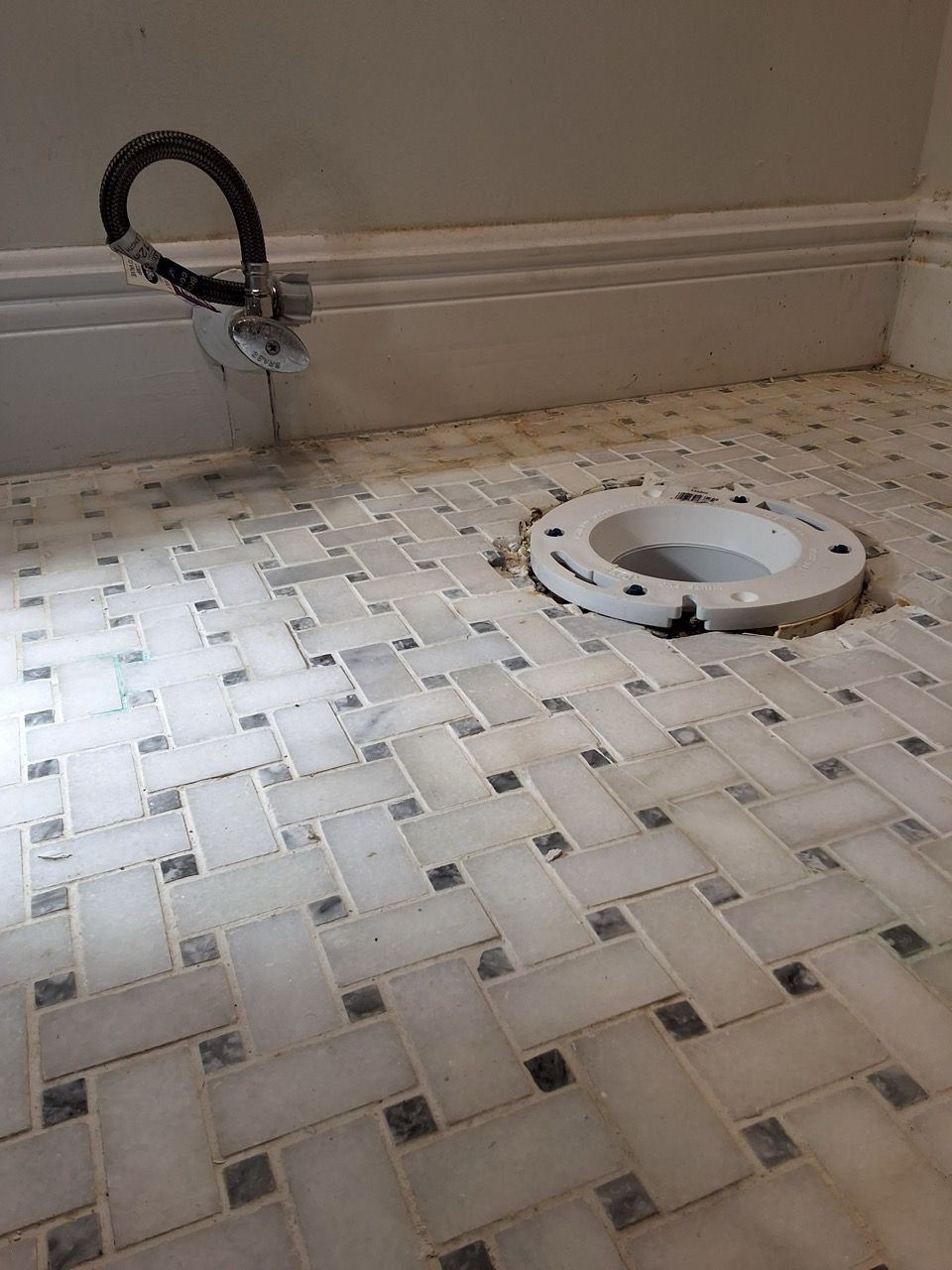 Bathroom floor with a toilet flange and a black hose near the wall, over a white-and-gray mosaic tile floor