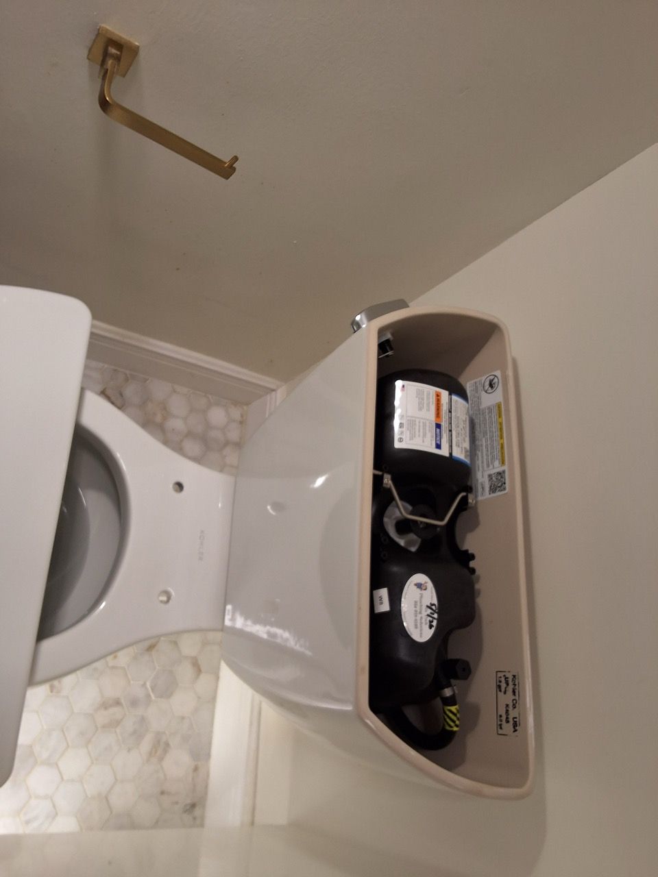 Open toilet tank showing internal flush mechanism and water supply valve, with the tank lid nearby.