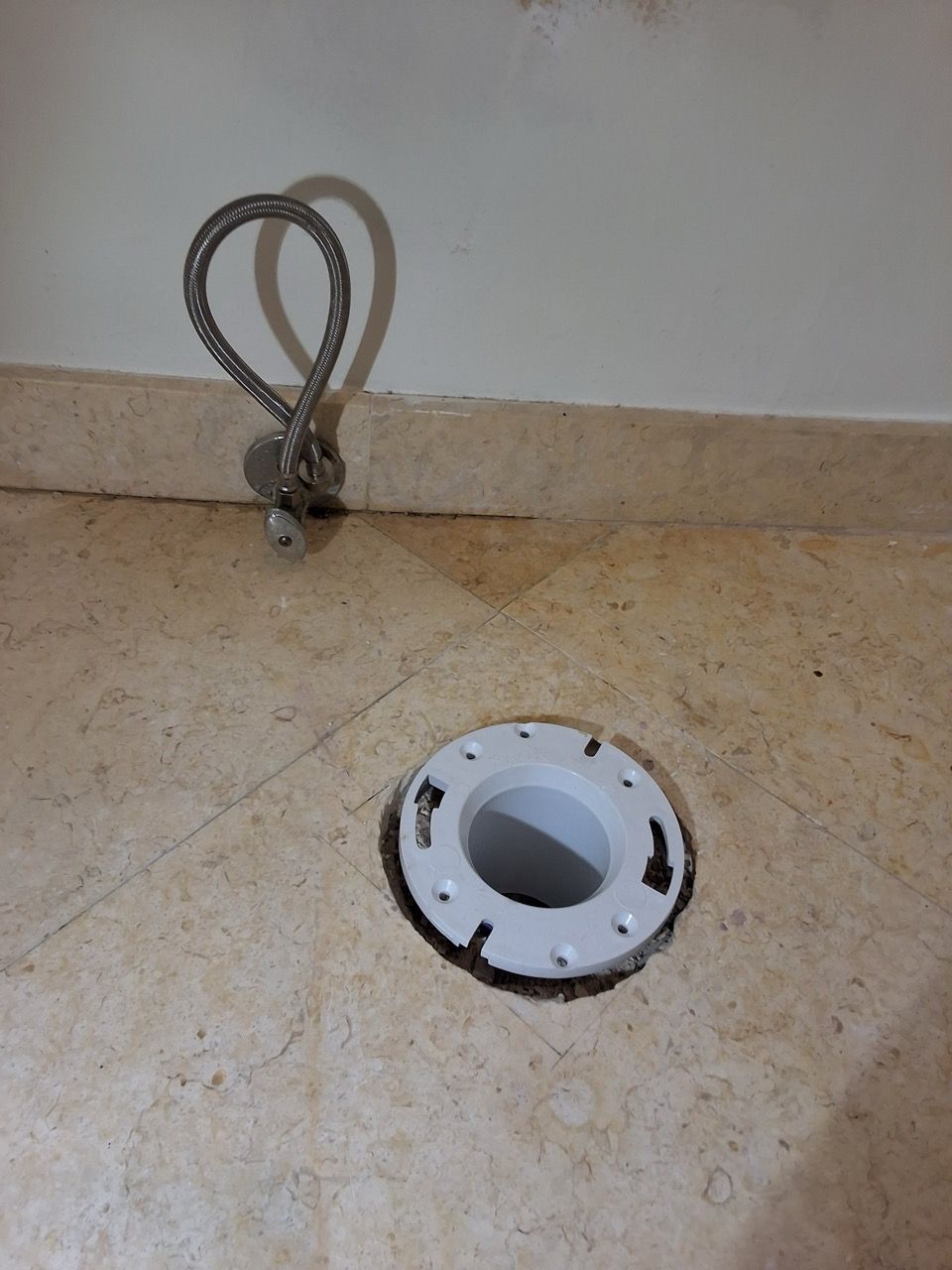 Floor drain opening in a tiled bathroom corner with a flexible hose at the wall.