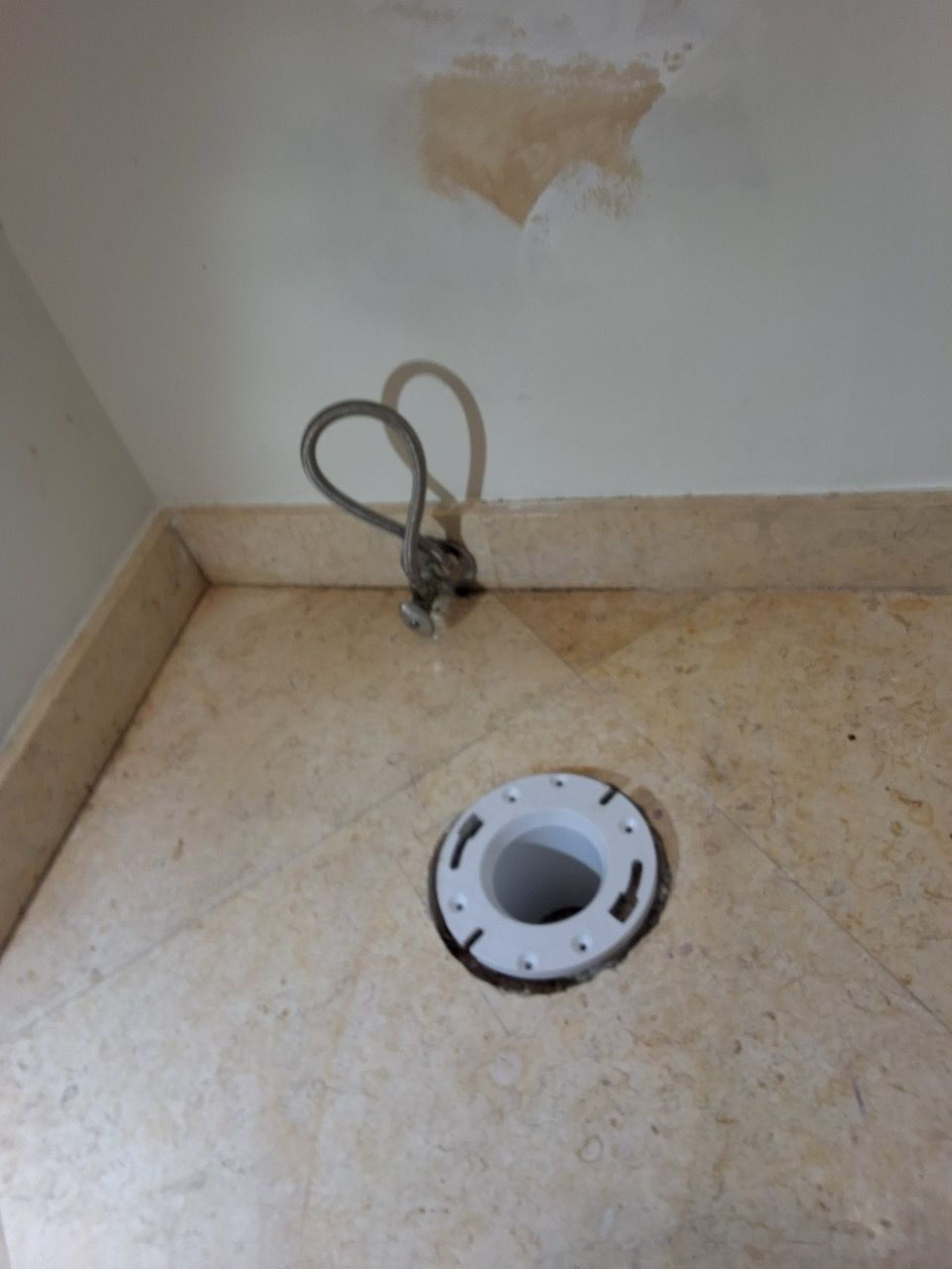 Bathroom floor with a toilet flange opening and exposed plumbing against a bare wall.