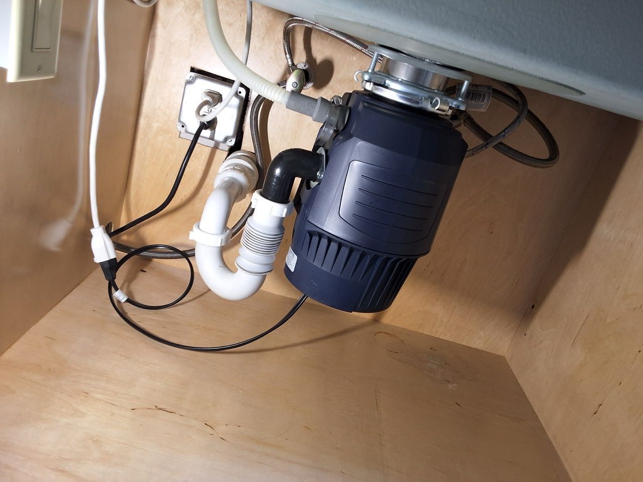 Under-sink garbage disposal and white plumbing pipes in a cabinet, with black cords and a blue container