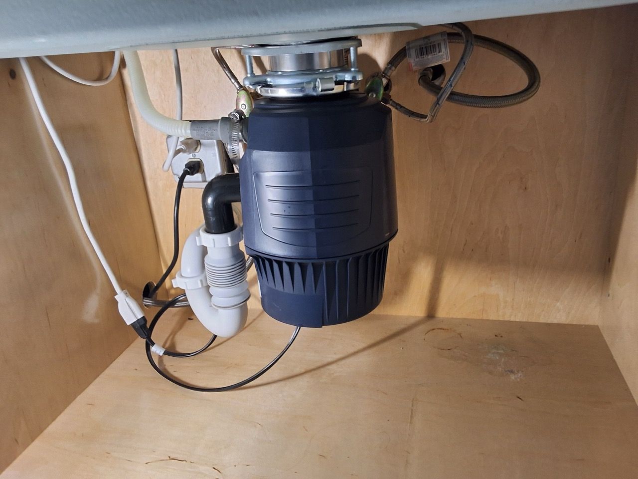 Under-sink garbage disposal and plumbing inside a cabinet with exposed wires and a power outlet