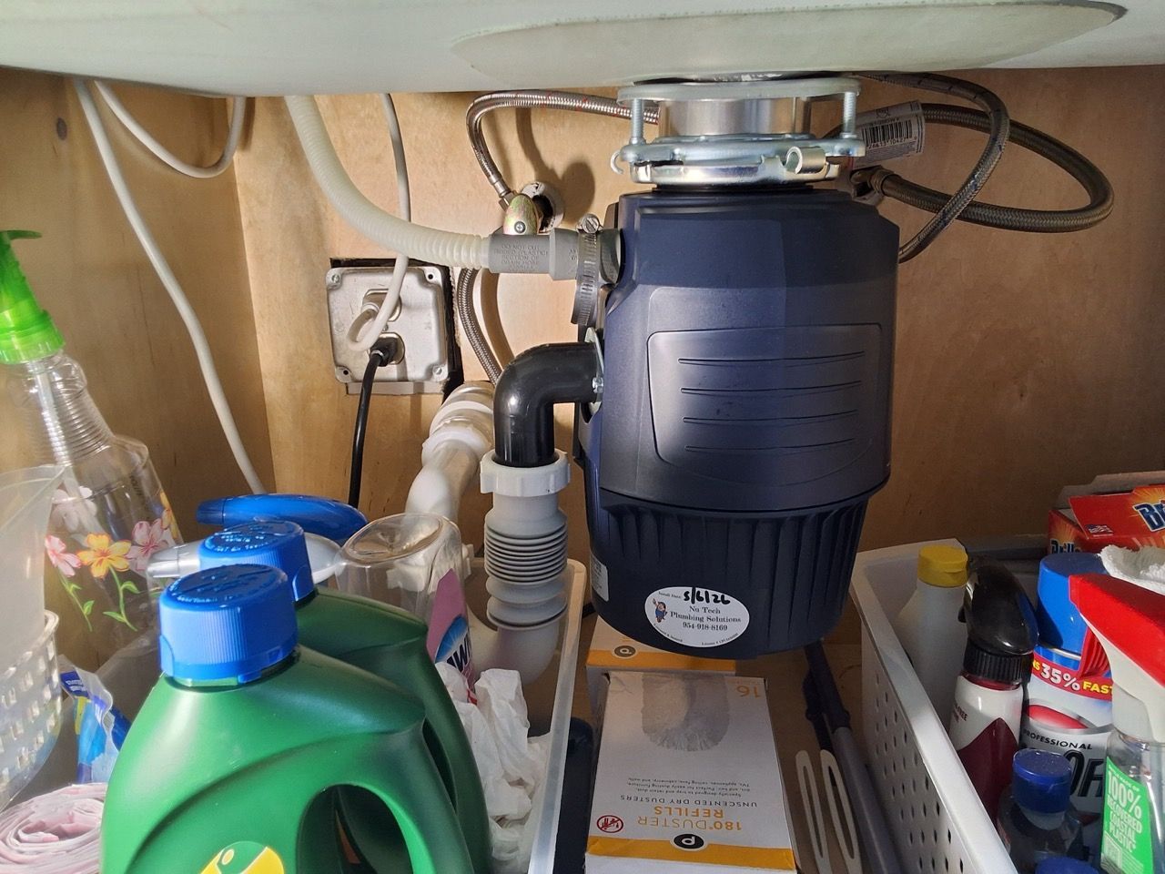 Under-sink cabinet with a dark garbage disposal, plumbing pipes, and cleaning supplies.