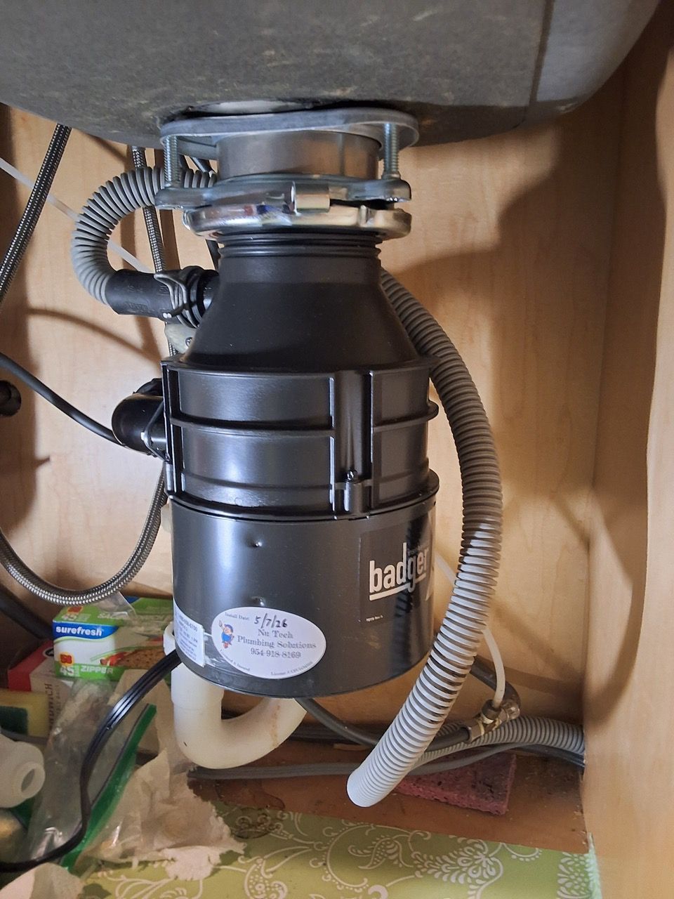 Black garbage disposal mounted under a sink with hoses and wiring in a cabinet.