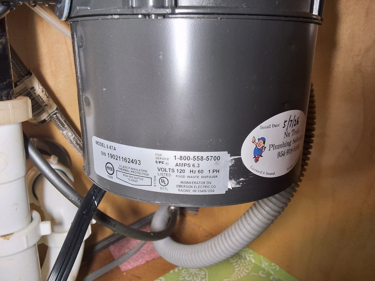Gray garbage disposal under a sink with white labels and hoses in a cabinet.