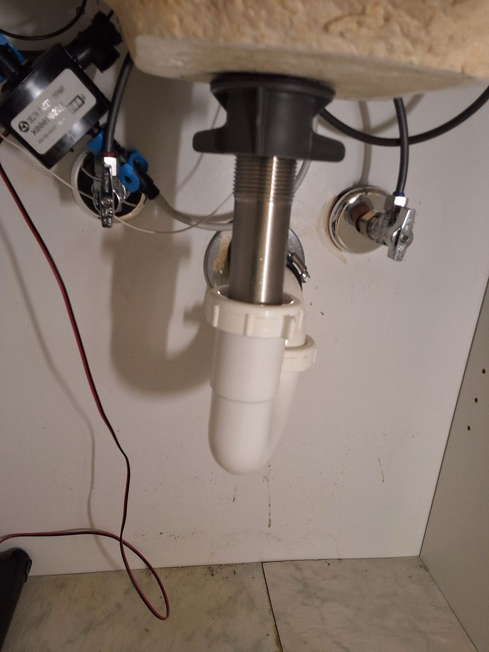 Under-sink plumbing with white drain pipe, metal sink drain, and exposed wires on cabinet wall