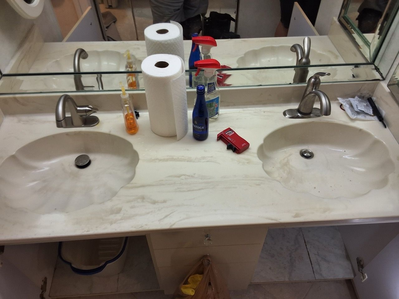 Double sink bathroom vanity with mirrors, soap dispensers, paper towels, and cleaning items on the counter