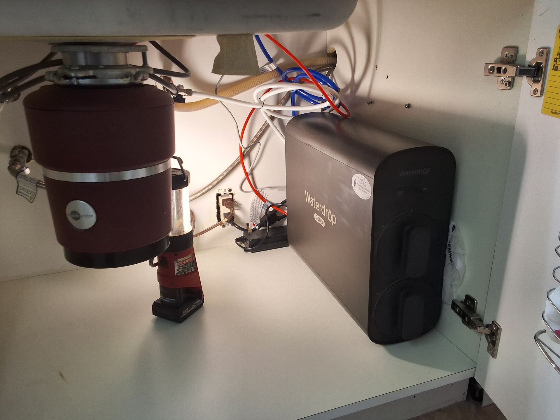Under-sink plumbing with a water filter canister, pipes, and a black appliance in a cabinet