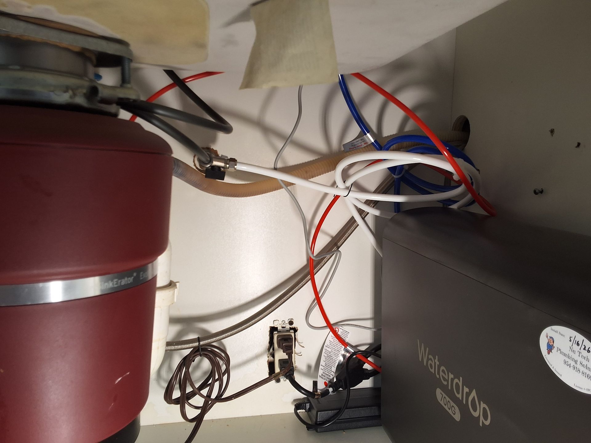 Wires and plumbing clutter the space beneath a sink or cabinet, with a red tank and gray box visible.