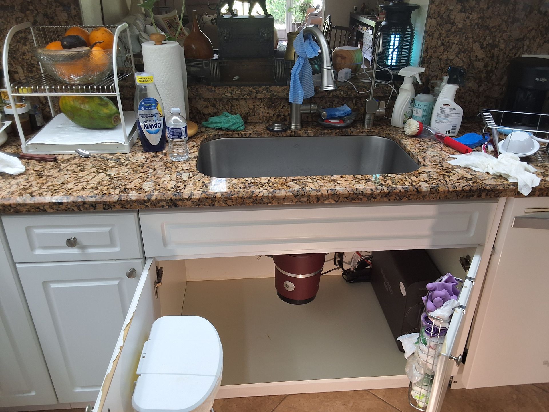 Kitchen sink with granite countertop, cluttered counter, white cabinets, and a fruit bowl by the window.