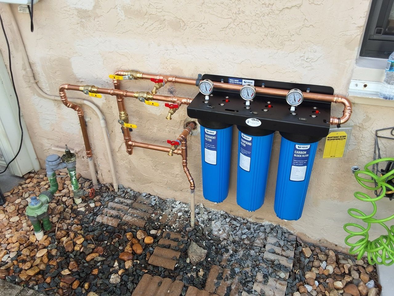 Blue water filters and copper pipes mounted on an exterior wall beside landscaping.