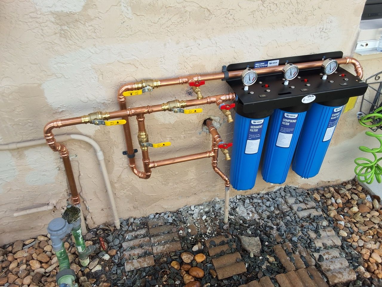 Water filtration system with blue canister filters and copper pipes mounted on an exterior wall.