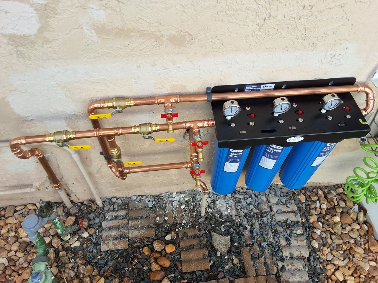 Outdoor water filtration system with copper pipes and three blue filter canisters mounted on a wall