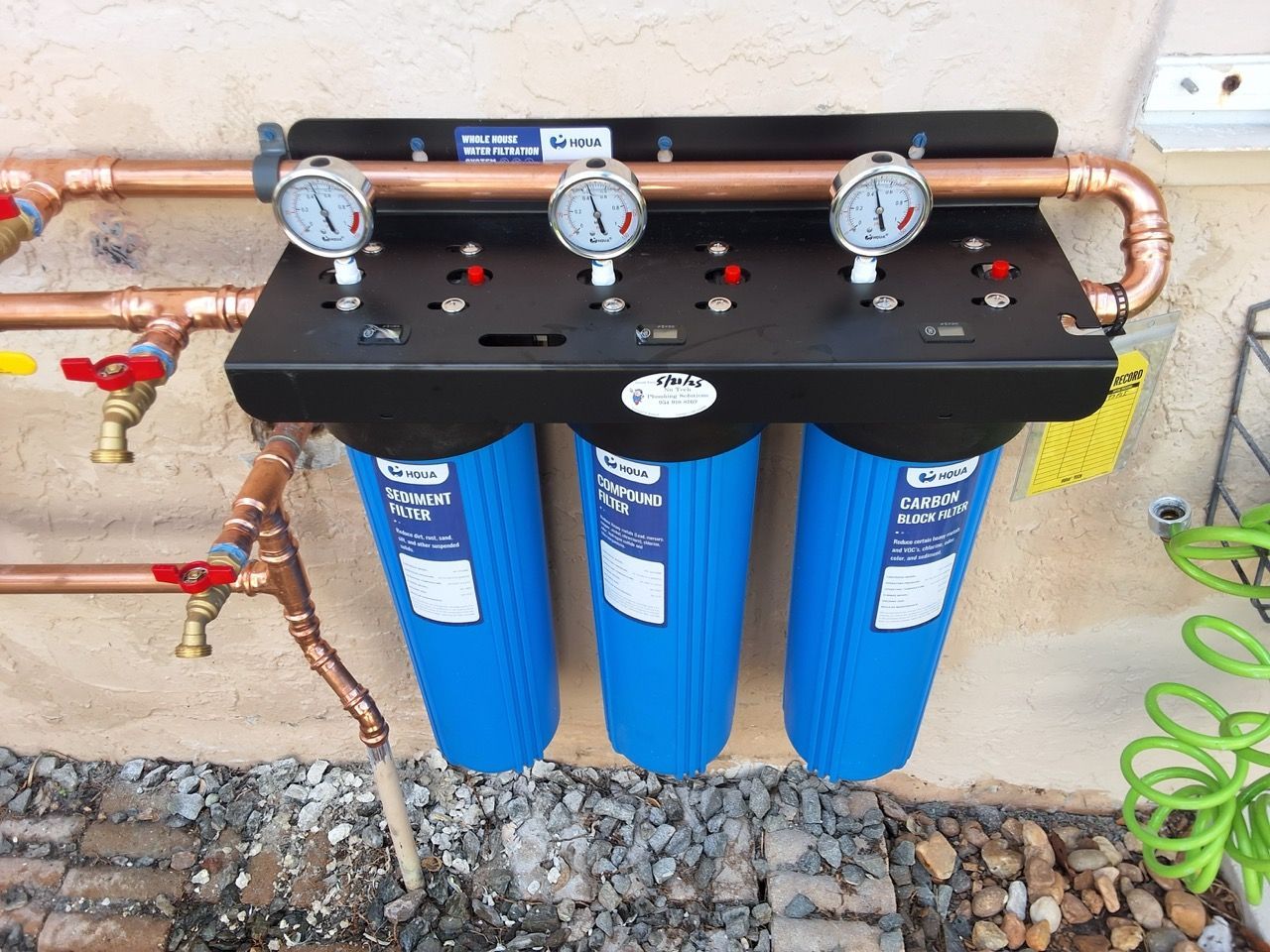 Three blue water filter housings with pressure gauges and copper pipes mounted on a wall outdoors