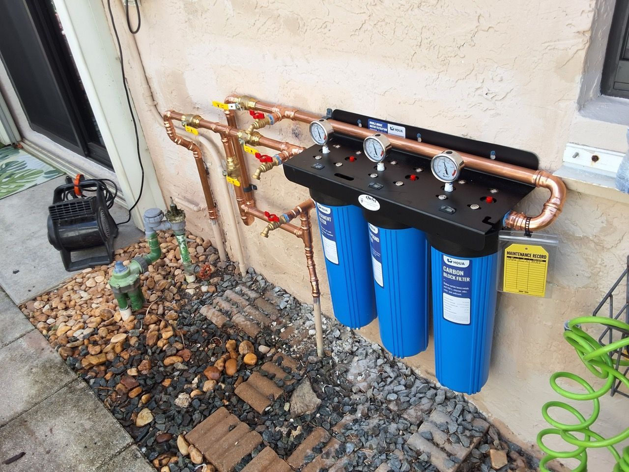 Blue water filter system mounted on exterior wall beside pipes, rocks, and a garden hose
