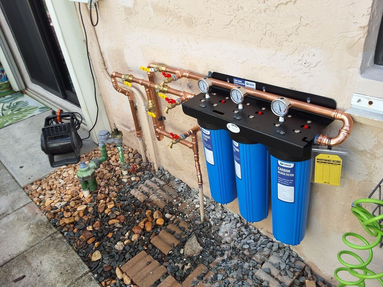 Water filtration unit with blue canisters mounted on an exterior wall beside a hose and gravel bed